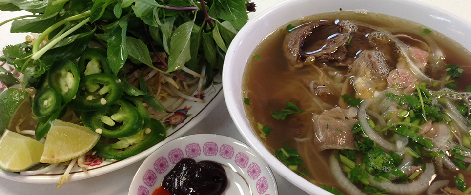Bowl of noodle soup with beef and herbs beside a plate of fresh greens, jalapeno slices, and lime wedges on a wooden table, warm and inviting mood
