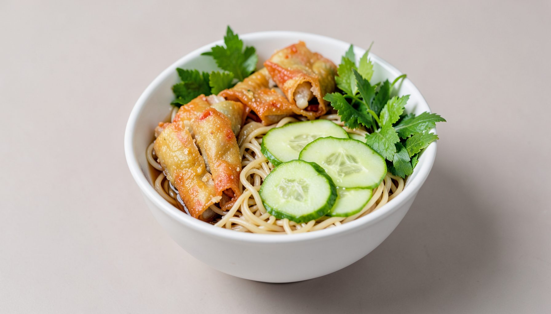 Vermicelli bowl with crispy spring rolls, cucumber slices, and bright green herbs on a neutral background